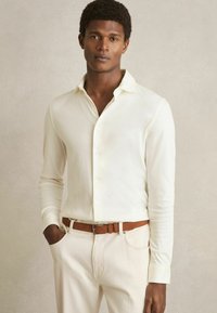 Cămașă albă cu mâneci lungi, textură netedă, guler clasic, completată cu o curea maro, purtată cu pantaloni bej deschis.