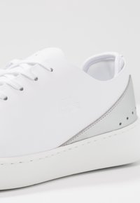 Zapatilla de cuero blanca con puntera redonda, detalle plateado en el talón y logo en relieve. Cuenta con cordones planos y suela de caucho texturizada.