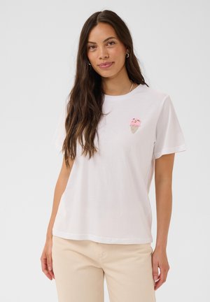 Ung kvinde med langt mørkt hår iført en hvid t-shirt med et lille pink isvaffel-design, beige bukser og sølv hoop-øreringe.