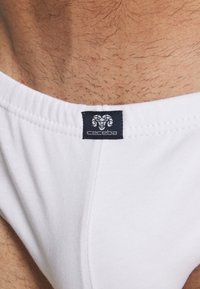 Weiße Baumwoll-Slip für Männer mit einem Logo-Tag, das ein Widderdesign zeigt. Der Stoff wirkt weich und glatt, mit einem elastischen Bund.