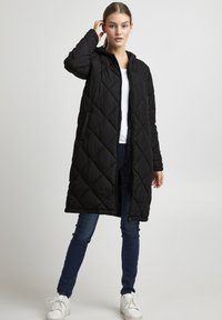 Cappotto lungo trapuntato nero con cappuccio, chiusura a zip e tasche laterali. Indossato sopra una camicia bianca e jeans blu, con sneakers bianche.