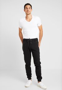 Zwarte joggers met een trekkoord in de taille, twee zakken met rits en een taps toelopende pasvorm, gecombineerd met een witte v-hals t-shirt en witte sneakers.