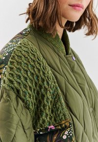 Prošívaná zelená bunda s texturovaným pleteným ramenem, květinovými vzory a žebrovaným límcem s fialovými kovovými detaily.