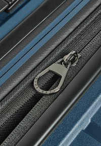 Primo piano di un tirante della cerniera nera su una valigia rigida blu con texture, che mostra i dettagli dei componenti in tessuto e metallo.