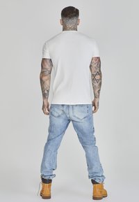 Tricou alb cu mânecă scurtă, cu dungi, asortat cu blugi de denim bleu deschis și cizme de lucru maro, evidențiind o croială confortabilă și un stil casual.