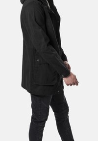 Giacca nera con cappuccio, dal taglio leggermente rilassato, dotata di bottoni a pressione, due tasche laterali e maniche testurizzate. Abbinata a pantaloni neri attillati.
