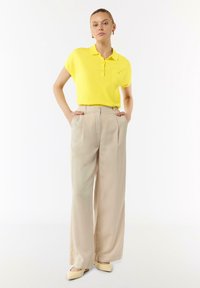 Gelbes Polo-Shirt mit kurzen Ärmeln und einer Brusttasche, kombiniert mit hellbeigen, weiten Hosen und blassen, gelben Schuhen.