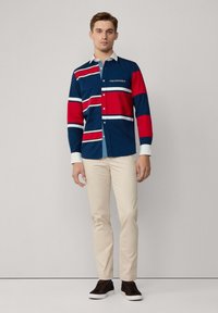 Camisa de manga larga para hombre en azul marino con rayas rojas y blancas, con un diseño de botonadura y cuello en contraste. Combinada con pantalones beiges.
