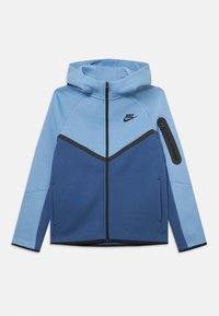 Nike bunda so striedavou kompozíciou v svetlom a tmavom modrom odtieni. Vyrobená z jemného materiálu, s vreckom na zips na rukáve a kapucňou.