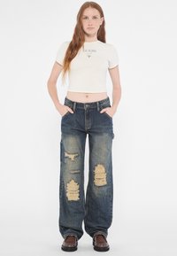 Camiseta corta blanca con el logotipo "GUESS"; pantalones cargo de mezclilla azul desgastados con grandes rasgaduras; zapatos marrones; modelo de pie con las manos en los bolsillos.