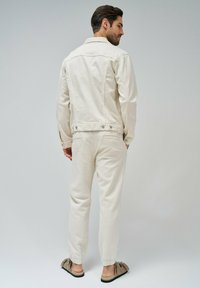 Chaqueta de mezclilla beige con cuello, bolsillos traseros y cierres de botón, combinada con pantalones ajustados a juego y zapatos cómodos de deslizar.