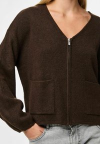 Brahns džemperis ar rāvējslēdzēju, V-veida kakla izgriezumu, garām piedurknēm, divām priekšējām kabatām un teksturētu audumu. Ikdienišķs stils, kas ir piemērots kārtošanai.