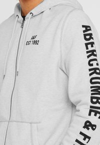 Grauer Kapuzenpullover mit Reißverschluss aus Baumwollmischung, mit Kordelzugkapuze, Vordertaschen und schwarzem Schriftzug auf dem Ärmel mit der Aufschrift "ABERCROMBIE & FITCH."