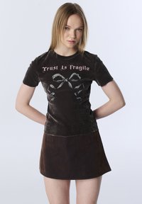 Donker fluwelen T-shirt met korte mouwen met de tekst "Trust Is Fragile" in roze en een afbeelding van een strik; gecombineerd met een donkerbruine A-lijn rok.