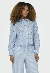 Camisa de botones azul claro con recortes de ojal, dobladillo con forma de scallop y mangas largas, combinada con pantalones con cordón de rayas claras.