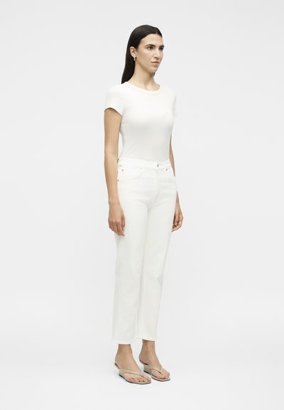 Γυναίκα στέκεται, φορώντας άσπρο εφαρμοστό τοπ με κοντά μανίκια, λευκό τζιν ψηλόμεσο με ίσια πόδια και μπεζ σανδάλια με επίπεδη σόλα.