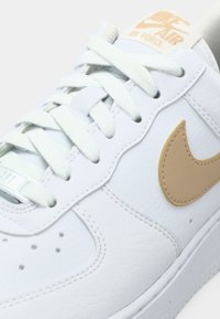 Białe skórzane sneakersy z jasnobeżowymi sznurowadłami, z złotym logo Nike i brązową etykietą na języku, na której widnieje napis "Nike Air Force".