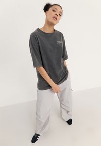 Übergroßes, dunkelgraues T-Shirt mit kurzen Ärmeln und einem bedruckten Design. Kombiniert mit hellgrauen Cargo-Hosen und schwarzen Sneakers.