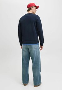 Dunkelblaues Sweatshirt mit Rundhalsausschnitt und geripptem Saum, kombiniert mit hellblauen weitem Jeans und einer roten Baseballmütze.