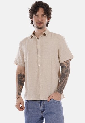 Hombre con cabello rizado que lleva una camisa beige de manga corta abotonada y jeans azules, mostrando tatuajes detallados en ambos antebrazos.