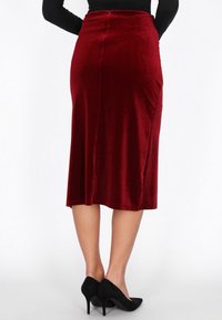 Pamutpiros bársony midi szoknya, testhezálló derékkal, oldalsó sliccel és sima textúrával, fekete magassarkú cipővel párosítva.