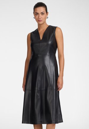 Femme aux cheveux foncés portant des boucles d'oreilles argentées et une robe midi en cuir noire sans manches avec un décolleté en V, debout devant un fond blanc.