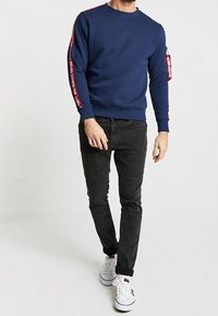 Marinblå sweatshirt med röda sidostråk, rund halsringning och ficka med dragkedja; ihop med svarta jeans och vita sneakers.