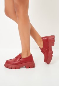 Czerwone skórzane buty na grubej platformie z cholewką w stylu mokasynów, z zakrzywionym szwem i detalem w postaci kokardy z przodu.
