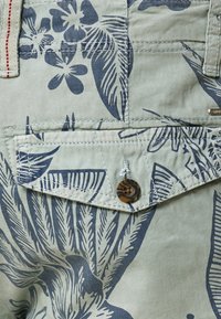Tissu vert clair avec un motif floral bleu foncé. Comprend une poche à rabat avec un bouton rond à rayures et des coutures contrastées.