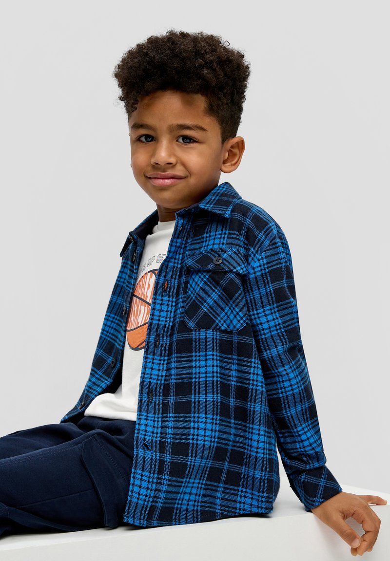 Karierter Hemd in Blau und Schwarz aus Flanell, mit einer Knopfverschluss-Front, einer Brusttasche und einer abgerundeten Saumlinie, getragen über einem weißen Grafik-T-Shirt.