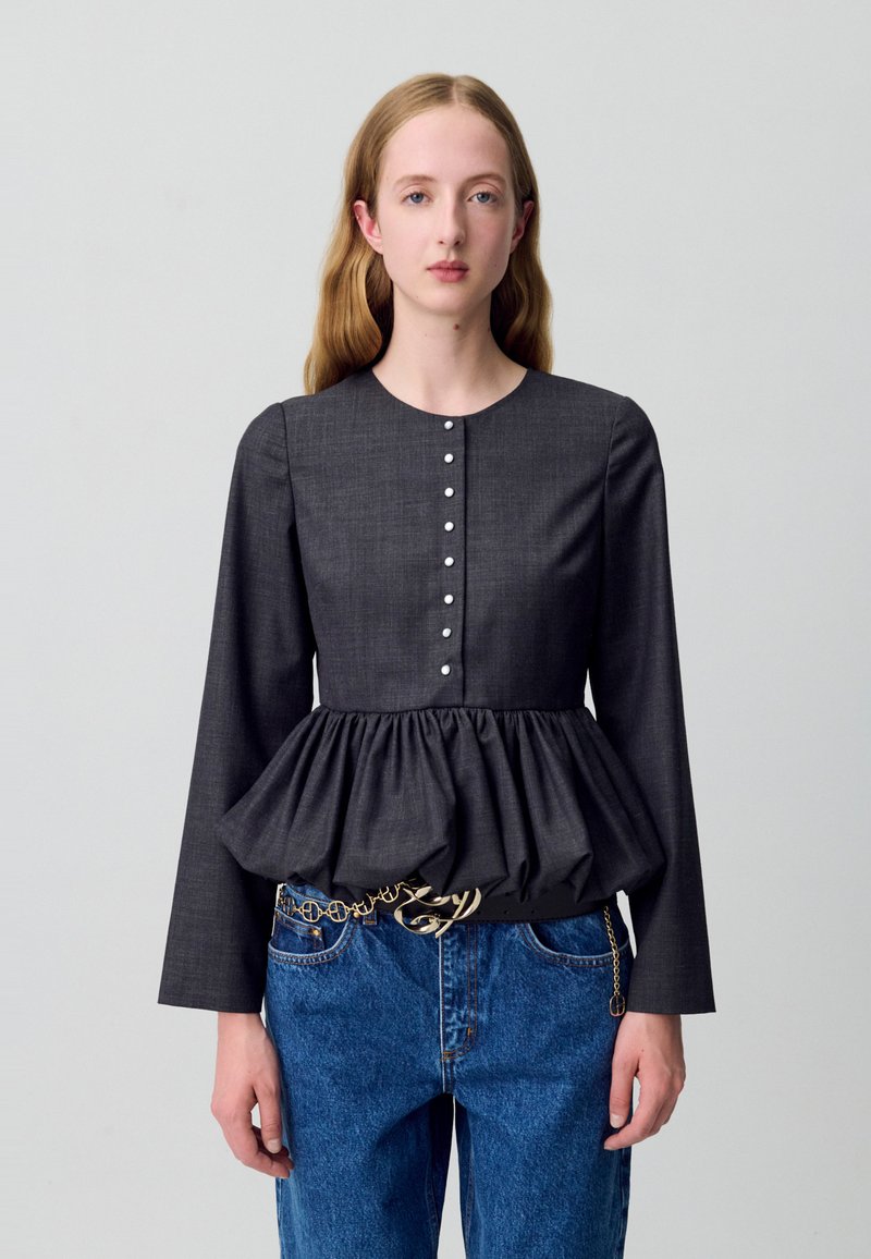 Top peplum grigio scuro con maniche lunghe, scollatura rotonda e dettaglio a bottoni sulla parte anteriore. Abbinato a jeans in denim blu e una cintura in catena dorata.