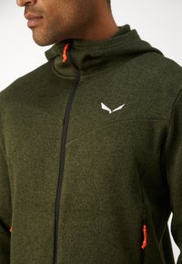 Άνδρας που φοράει ελιά πράσινη μπλούζα με φερμουάρ και μαύρο φερμουάρ με πορτοκαλί κορδόνια, με λευκό λογότυπο σε σχήμα πουλιού στο στήθος.