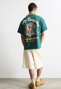 Homme de dos portant un T-shirt teal avec un graphique coloré de camion de tacos, ainsi que des shorts beiges et des bottes, sur un fond blanc uni.