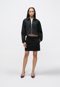 Prirezana črna jakna z zadrgo, kombinirana s črno krilo z logotipnim pasom. Model nosi bordo čeveljce z ostrim koncem.