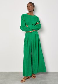 Groene twee-delige outfit met een lange mouwen top en wijde broek, gemaakt van gestructureerde stof. Geaccentueerd met een lichtgekleurde riem en sandalen.