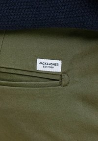 Maslinasta zelena tkanina s teksturiranim tamnoplave boje, s bijelom etiketom na kojoj piše "JACK & JONES EST. 1990" pričvršćenom sa strane dizajna.