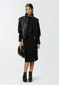 Giacca senza maniche in pelle nera, blusa nera aderente, gonna nera al ginocchio, borsa nera lucida e stivali alla caviglia brillanti con accenti dorati.