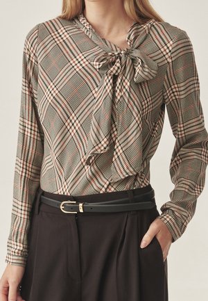 Vrouw die een beige geruit blouse met lange mouwen draagt, met een strik bij de hals, een zwarte riem en donkerbruine broek met hoge taille.