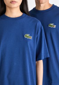 Camisetas de algodón azules con cuello redondo, que cuentan con un parche de logo de cocodrilo verde en el lado izquierdo del pecho. Ambas camisetas tienen mangas cortas.
