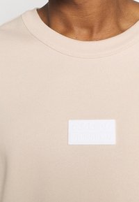 Beige sweatshirt with a round neckline, featuring a textured, white rectangular patch displaying the Adidas logo. Soft fabric, minimal design.