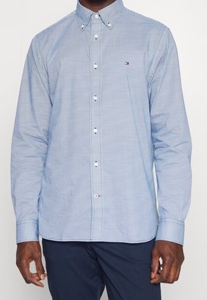 Mężczyzna w jasnoniebieskiej, wzorzystej koszuli z długimi rękawami i granatowych spodniach, stojący z ramionami swobodnie opuszczonymi wzdłuż ciała.