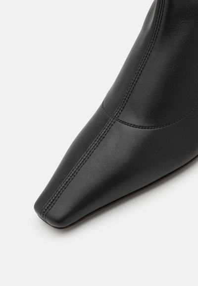 Ghete din piele neagră până la gleznă, cu vârf ascuțit, textură netedă și cusături vizibile pe laturi.