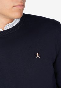 Maglione lavorato a maglia di colore blu navy con collo rotondo e polsini a costine. Presenta un piccolo logo ricamato di colore chiaro sul petto.