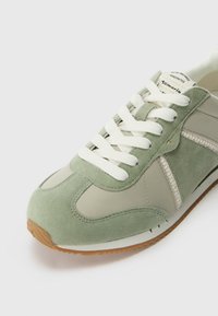 Sneaker verde in suede e sintetico con lacci bianchi, accenti tonali e suola in gomma. Presenta un pannello laterale con motivo e dettagli cuciti.