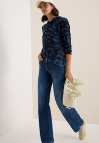 Navyblauwe hoodie met een patroon, blauwe jeans met een rechte snit, beige tas en lichte sneakers. De hoodie heeft een casual pasvorm en een capuchon.