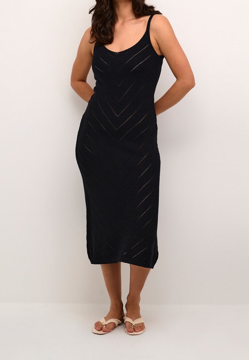 Femme portant une robe mi-longue noire en maille sans manches avec des motifs en chevrons, debout pieds nus sur un sol blanc contre un mur clair uni.