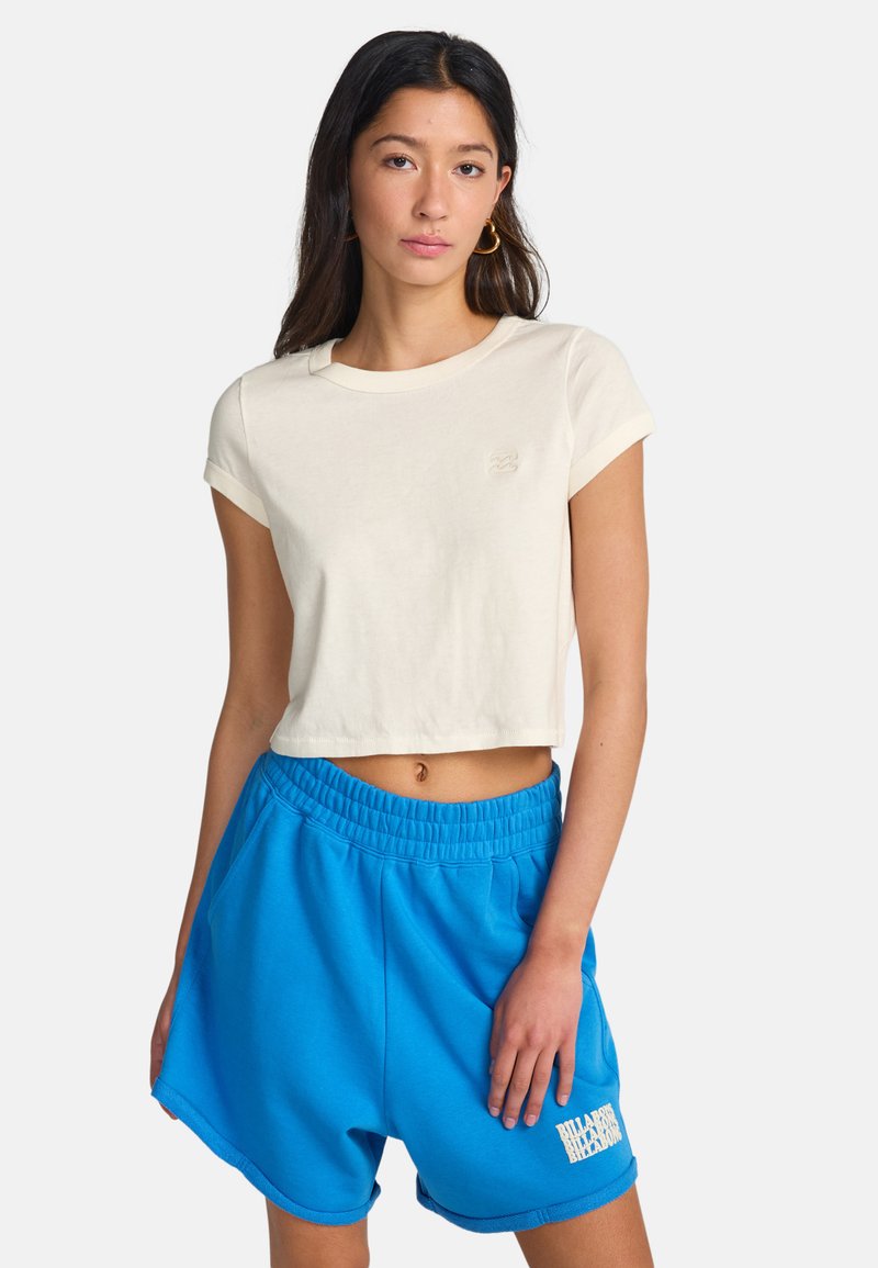 Nuori nainen, jolla on yllään kermaisen värinen lyhyt t-paita ja kirkkaan siniset, joustovyötäröiset shortsit, joissa on logo vasemmassa lahkeessa, seisoo valkoista taustaa vasten.