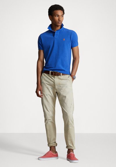 Camisa polo azul real de manga corta, pantalones caqui con manchas de pintura multicolores y zapatillas de lona rosas. Cinturón de cuero marrón.