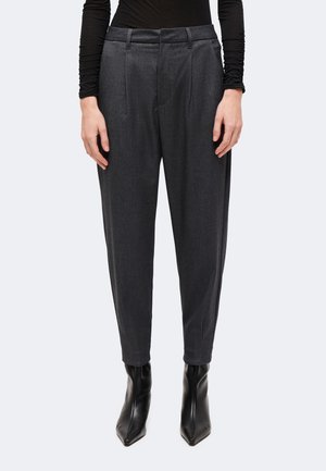 Personne portant un pantalon gris cintré avec plis, un haut noir moulant à manches longues et des bottines noires pointues, debout sur un fond uni.