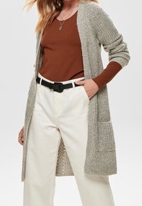 Vrouw in een bruine lange mouwen top, beige gebreide cardigan met zakken, witte hoge taillebroek en zwarte riem, met handen in de zakken.